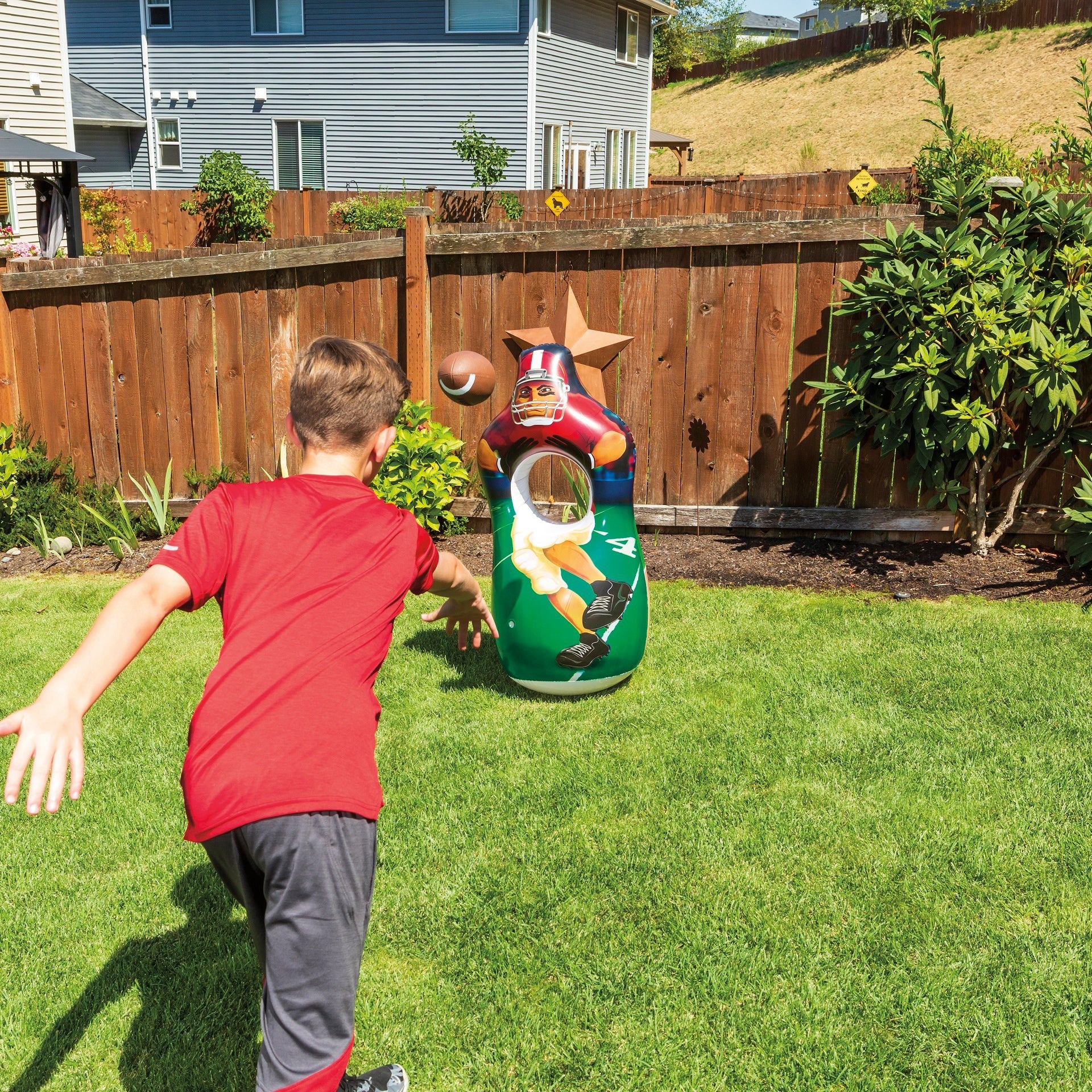 A boy in a red shirt throws a football toward the GO! Play Inflatable Toss by GO! Play, featuring a football player design, set up on grass in a backyard bordered by a wooden fence and houses.