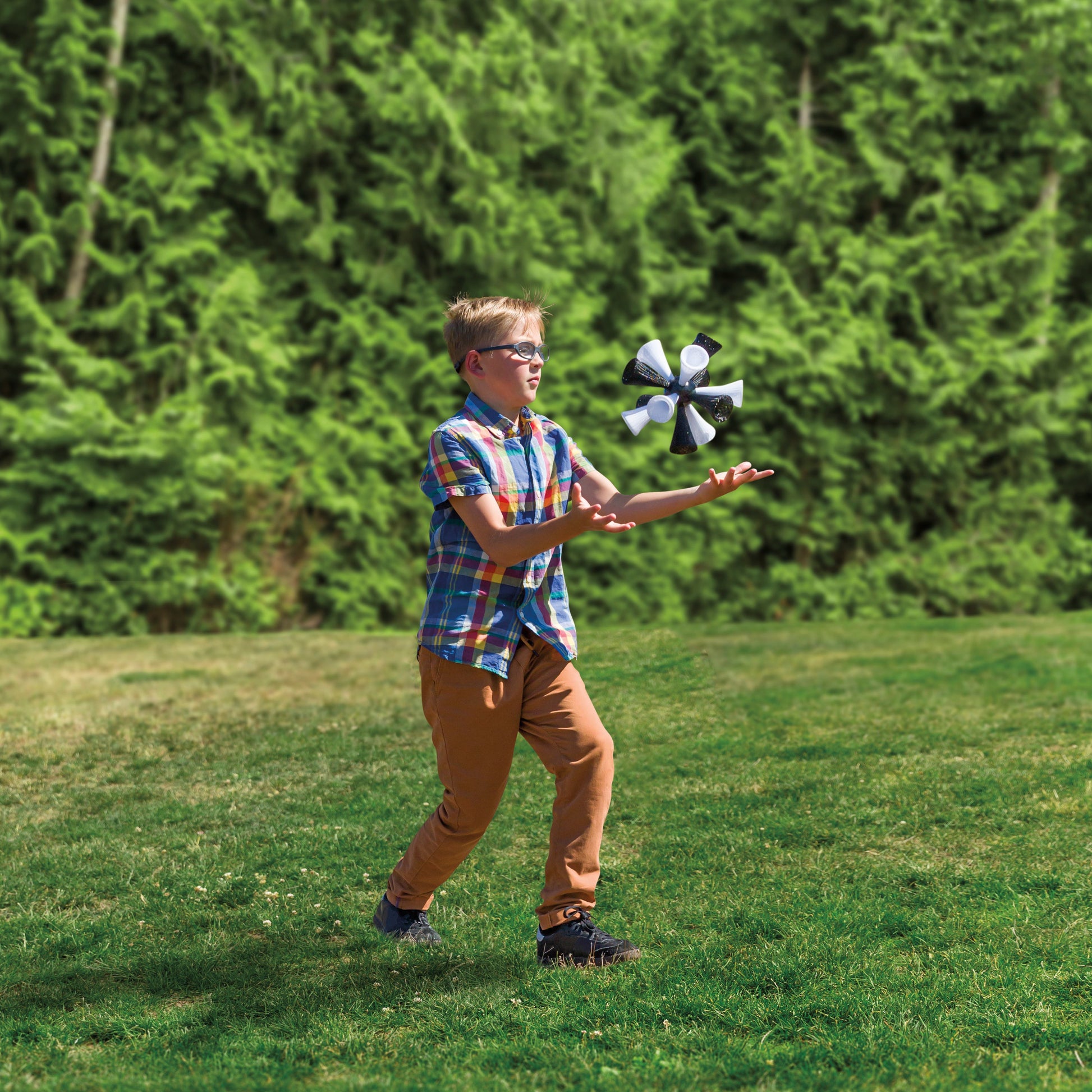 A boy in glasses, a plaid shirt, and tan pants plays outdoors on grass, reaching up to catch the GO! Play Jumbo Bash Birdie in the backyard with tall green trees behind him.