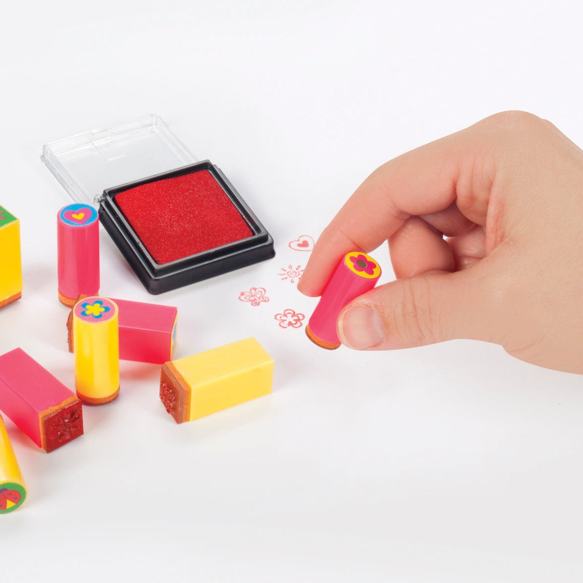 A hand uses a Toysmith Mini Stamp Set with a red ink pad to stamp colorful flower shapes on white paper, while other vibrant Toysmith Mini Stamp Sets and the ink pad inspire more creative designs nearby.