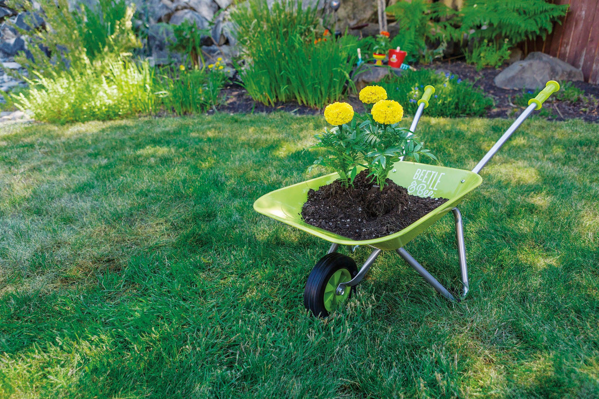 The Beetle & Bee Garden Kids Wheelbarrow by Beetle & Bee Garden, filled with soil and yellow flowers, sits on a grassy lawn with a "Bee the best" sign beside it and lush green plants and rocks in the background.