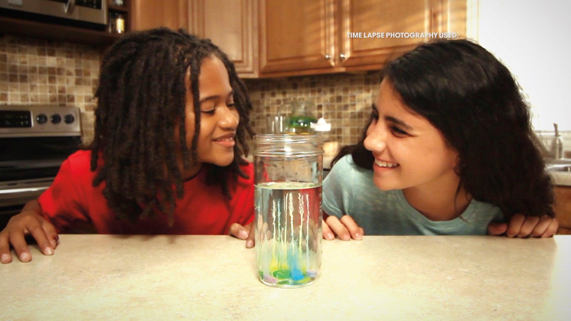 Two children smile at each other across a kitchen counter, with The Original Magic Rocks Crystal Growing Kit by NSI in a glass jar between them. Behind them are wooden cabinets and a tiled backsplash.