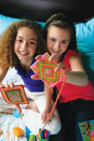 Two smiling young girls sit on a bed, proudly holding up colorful creations they made with the 4M-Craft Easy To Do Cross Stitch kit by 4M-Craft, showing off bright patterns and designs.