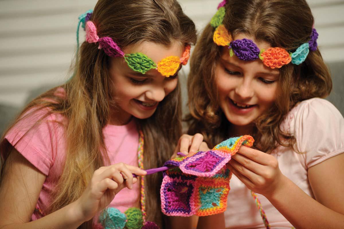 Two smiling girls use the 4M-Craft Easy To Do Crochet kit, working together on a colorful patchwork project. One holds a crochet hook while they both focus excitedly on their vibrant creation from 4M-Craft.