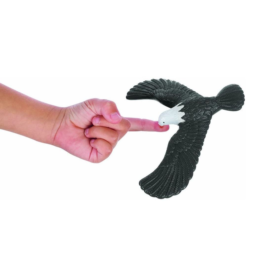 A hand’s outstretched finger balances the Toysmith Balancing Eagle by Toysmith on its beak, creating the illusion that the bird is flying past the hand against a white background.