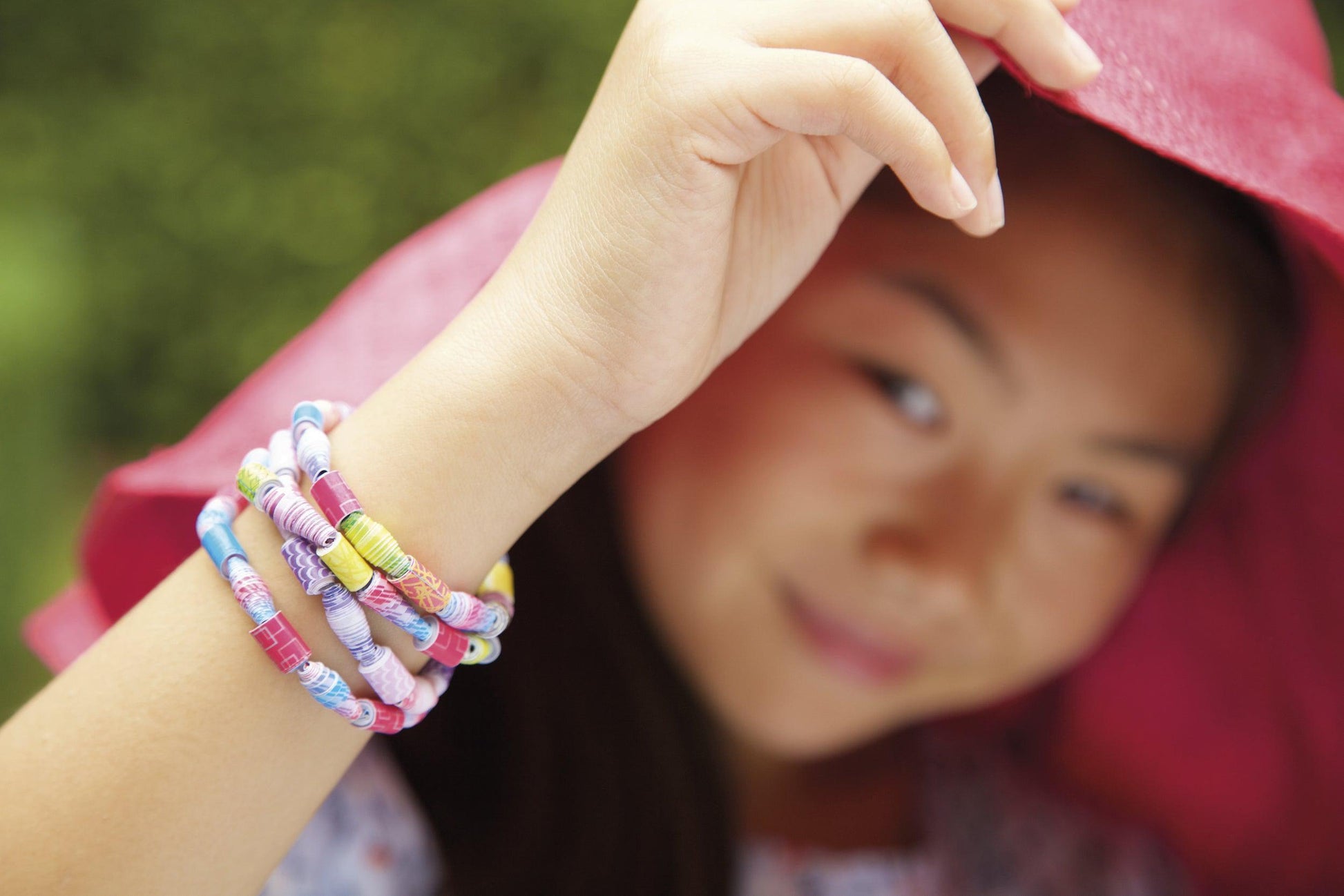 Wearing a large pink hat and several 4M-Green Creativity Recycled Paper Beads bracelets, a person smiles softly with a raised hand near their face against a backdrop of blurred greenery.