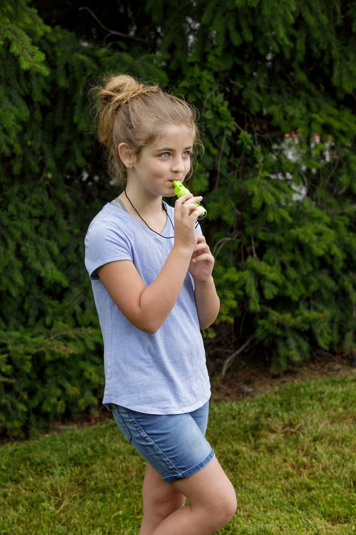 A young girl with light brown hair in a messy bun stands on grass near bushes, wearing a light blue t-shirt and denim shorts, sipping from a green juice pouch and holding her favorite Outdoor Discovery Survival Compass by Outdoor Discovery.