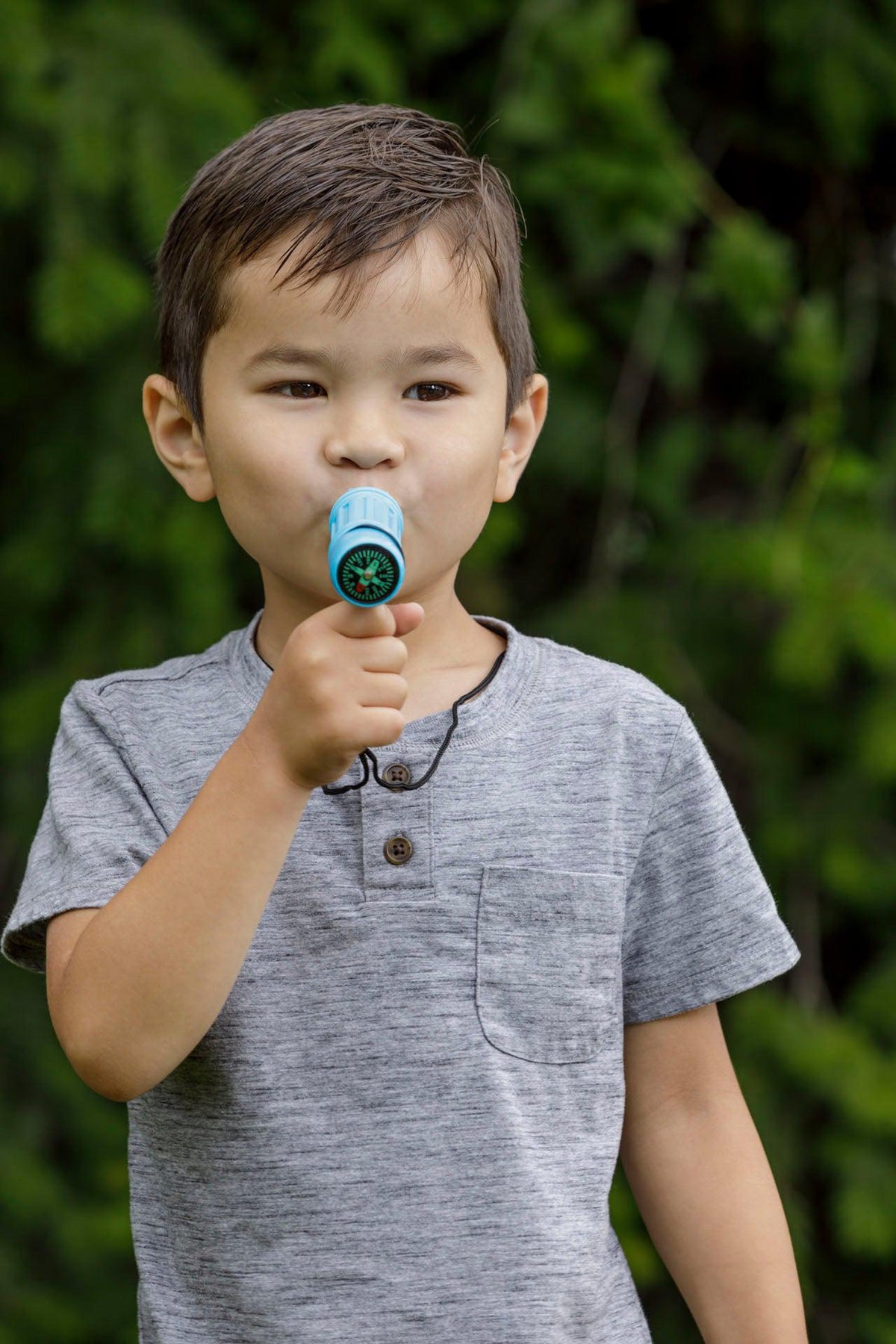 A young boy in a gray shirt stands outdoors, holding the Outdoor Discovery Survival Compass by Outdoor Discovery to his mouth as if speaking into it, with blurred green foliage in the background.