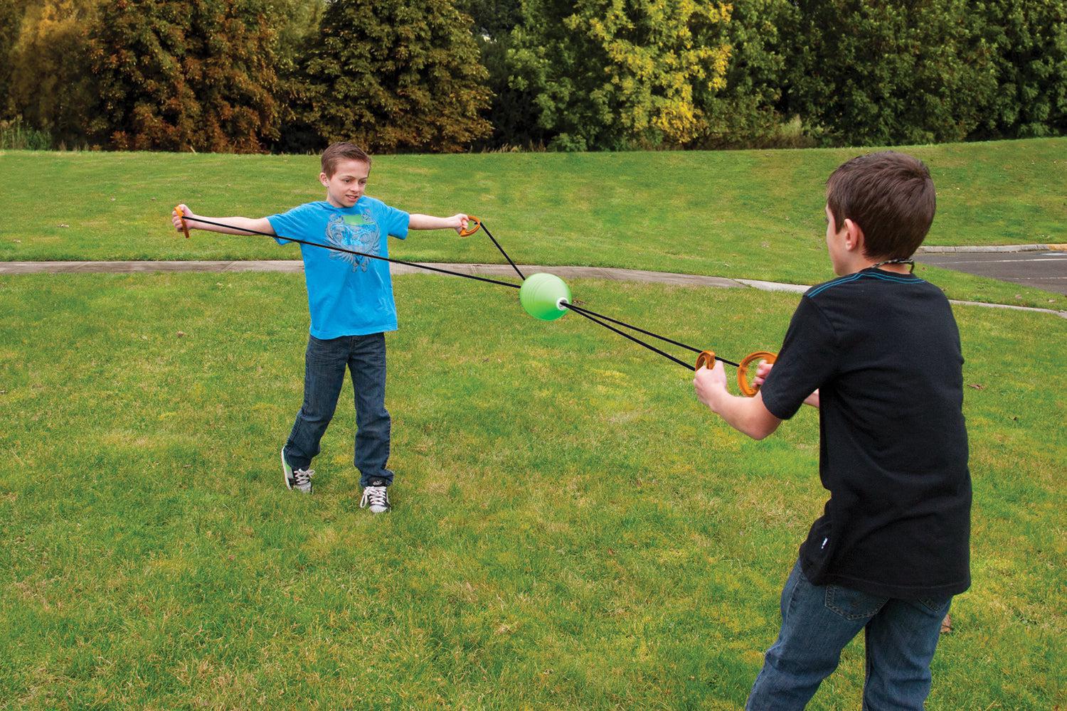Two boys play with the GO! Play Zip Ball by GO! Play, standing on grass and sliding a green ball back and forth using handles attached to strings. Trees in the background make it perfect for backyard or park fun.