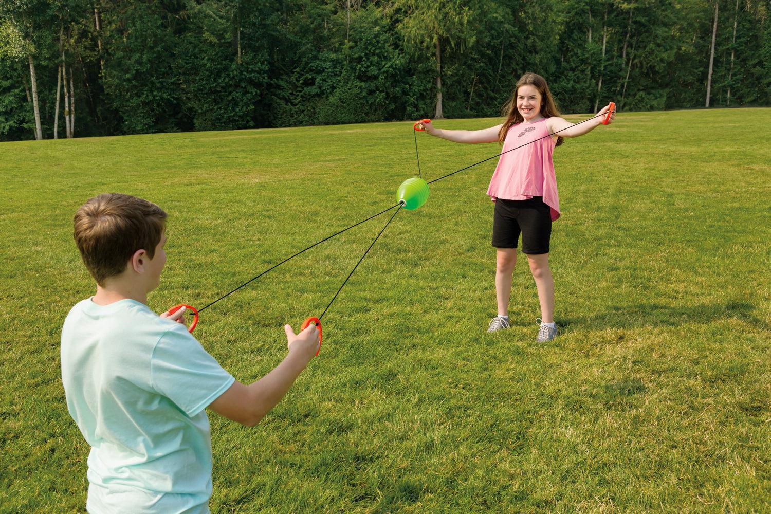 Two kids play outdoors with the GO! Play Zip Ball by GO! Play, pulling handles attached to strings as a green ball zips between them—perfect for backyard or park fun with trees in the background.