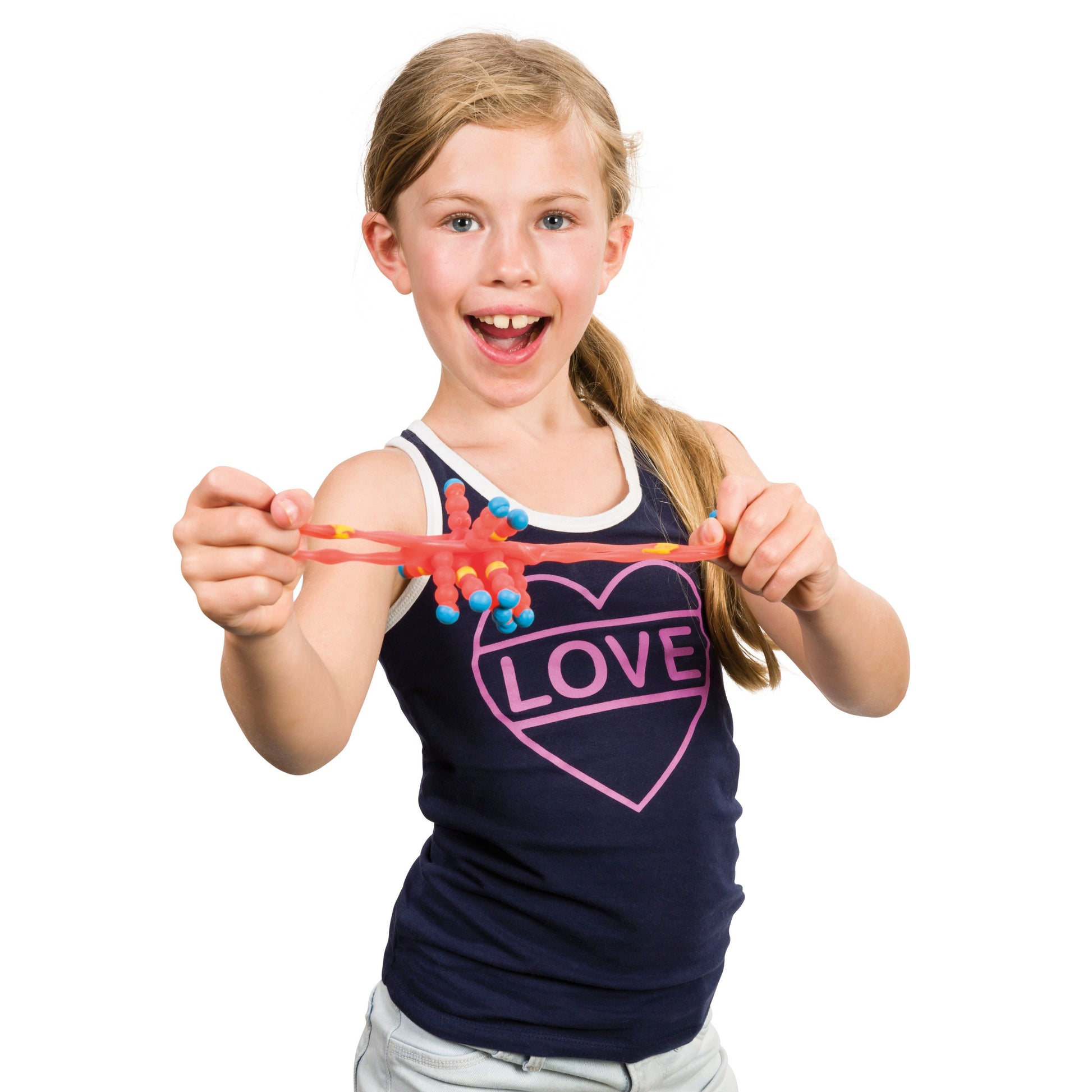 A smiling young girl with blonde hair stretches the Toysmith Colorful Atom Ball. She wears a navy blue tank top with a pink heart and “LOVE” on it, and light shorts, standing against a white background. Brand: Toysmith.
