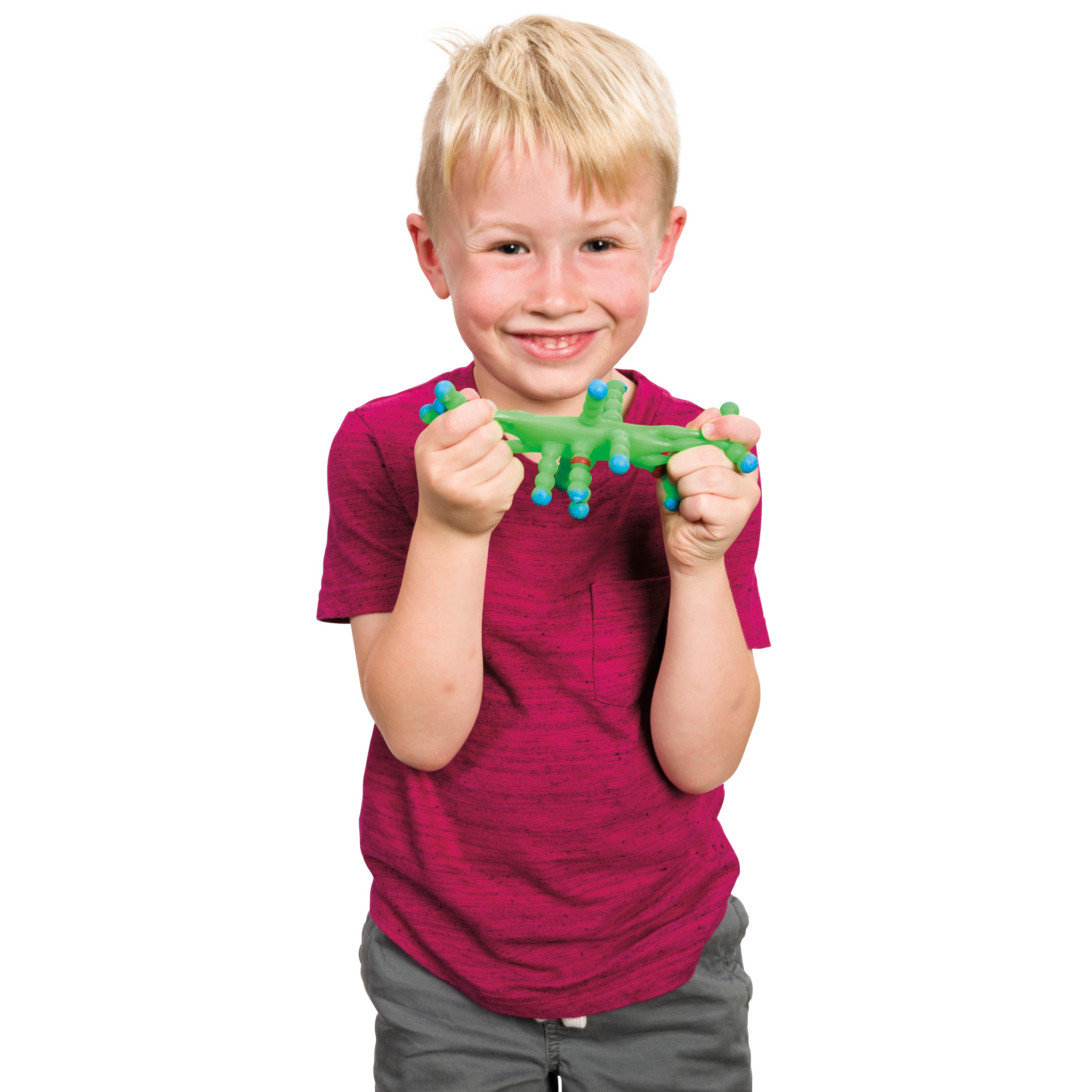 A smiling young boy with blond hair, wearing a pink shirt and gray pants, holds the Toysmith Colorful Atom Ball by Toysmith—a green stretchy toy with blue and purple suction cups—against a white background.