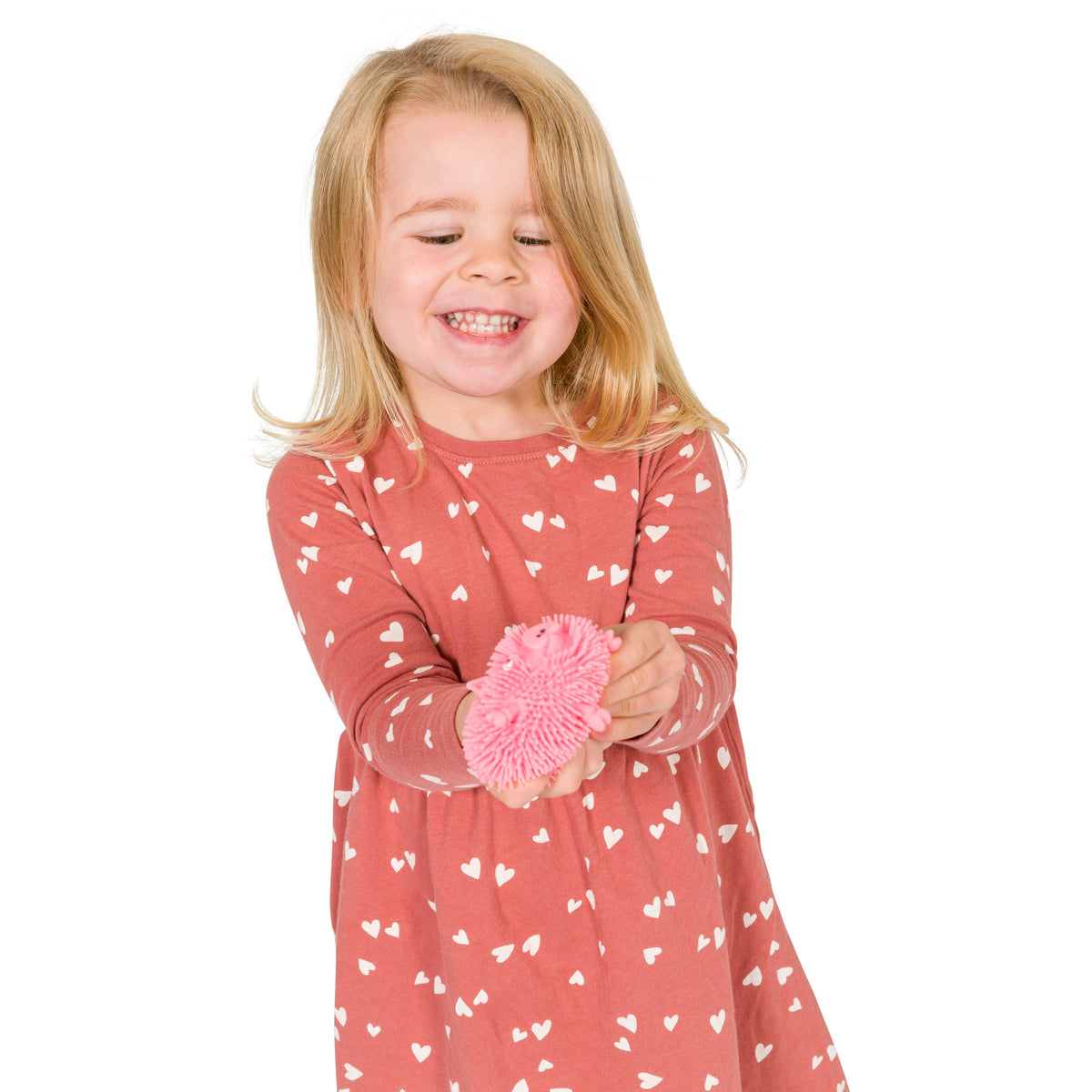 A young blonde girl in a pink dress with white hearts smiles and holds out a pink Farm Fresh Wee Critter Puffs sensory toy against a white background.