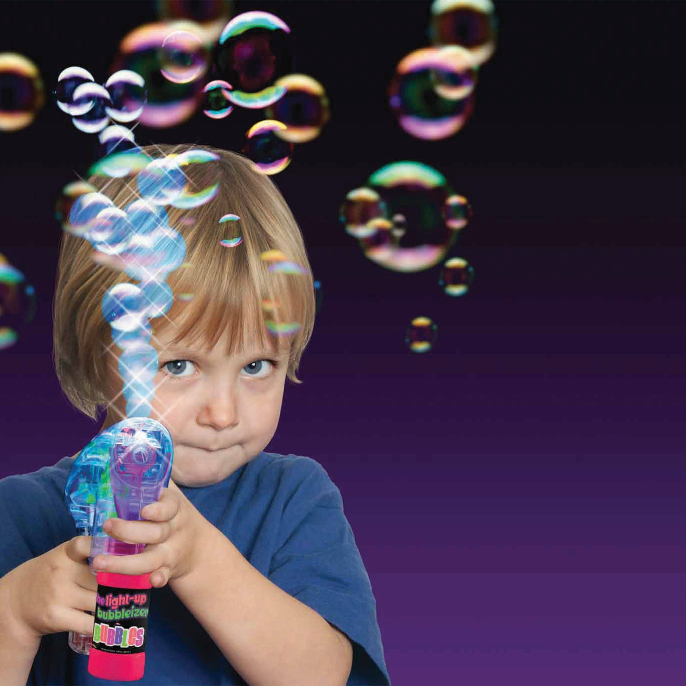 A young child in a blue shirt uses the We're Always Thinking Light Up Bubbleizer by We're Always Thinking, creating bubbles that float against a dark purple background while looking intently at the camera.