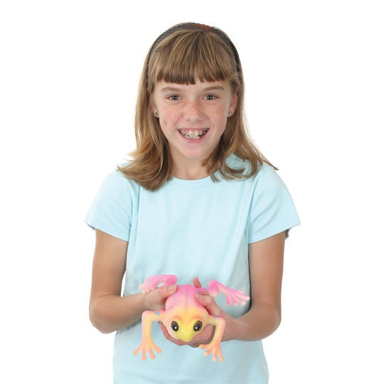 A smiling young girl with braces and light brown hair, in a light blue shirt, holds a Toysmith Ginormous Grow Frog—a large pink and yellow expanding frog toy by Toysmith—against a white background.