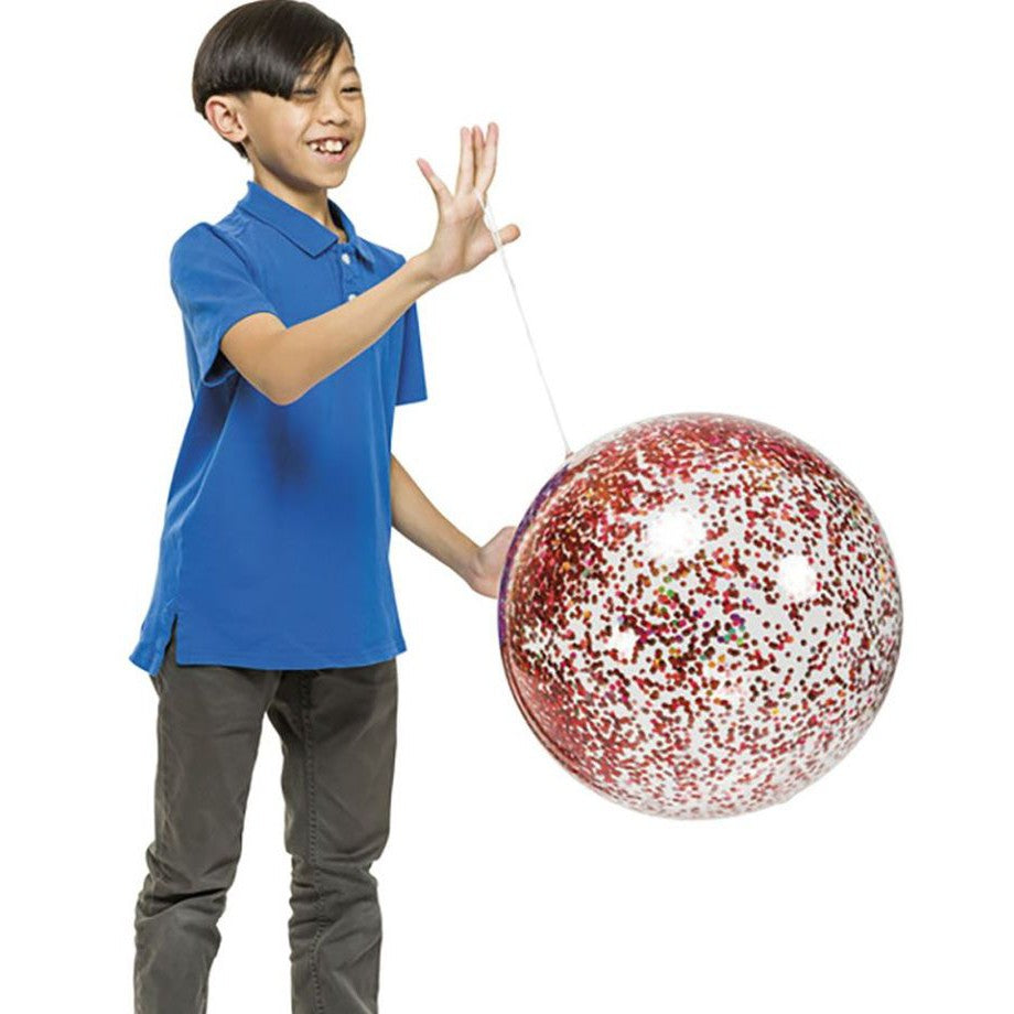 A smiling child in a blue polo and gray pants holds a large YAY! Glitter Punch Balloon by YAY!, filled with red, white, and pink confetti, featuring an elastic band for active play.