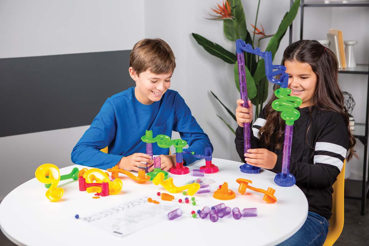 Two children sit at a white table assembling the Toysmith 80pc Marble Run building set, featuring colorful plastic pieces and marbles. An instruction sheet is on the table, with a tall plant visible in the background.