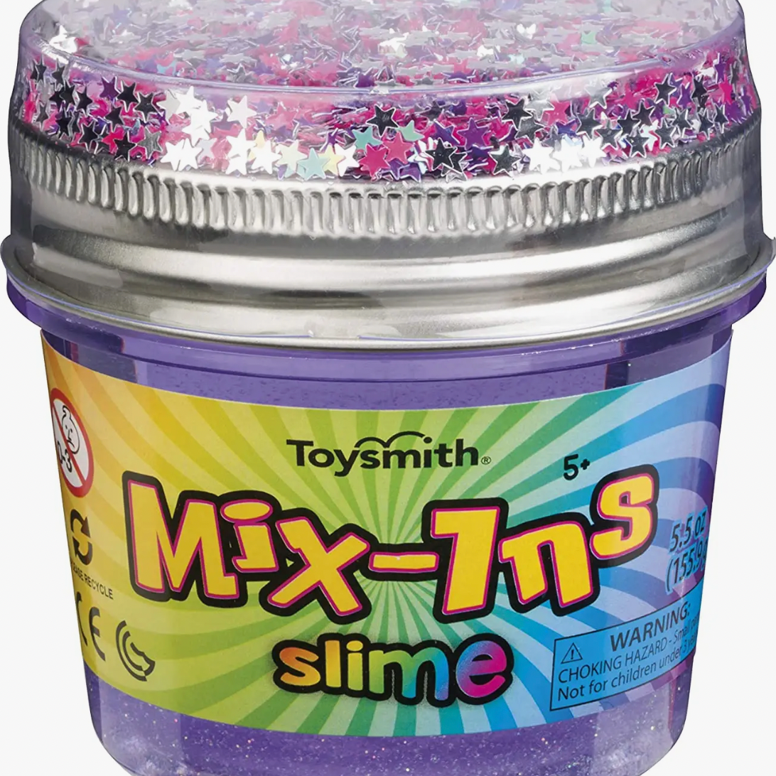 A Toysmith Mix-Ins Slime Asst container with a purple base, clear lid, and rainbow label holds star-shaped glitter. Bright Toysmith packaging shows off the playful mix-ins inside this slime toy.
